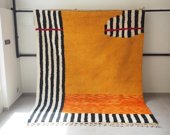 May include: A large, rectangular rug with a vibrant color palette. The rug features a mustard yellow central field, bordered by black and white striped sections. Accents include an orange band and a black and white striped design with a red stripe.