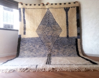 Op de afbeelding: Een wit en blauw Marokkaans tapijt met een geometrisch ruitpatroon. Het tapijt heeft een franje rand en is gemaakt van wol.