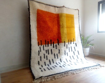 May include: A large, rectangular rug with a cream-colored background. The rug features a bold geometric design with orange and yellow blocks, and black vertical dashes. The edges are trimmed with a black border and fringe.