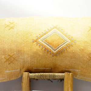 May include: A rectangular, yellow cushion with an embroidered diamond pattern in the centre. The cushion has a textured surface and is adorned with geometric designs in white and gold. The cushion is resting on a wooden frame.