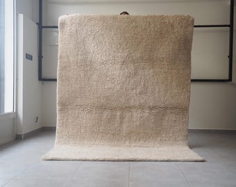 Puede incluir: Una alfombra beige rectangular grande con un pelo grueso y texturizado. La alfombra se exhibe en interiores, con una parte doblada. El color neutro y la textura afelpada sugieren un enfoque en la comodidad y la calidez.
