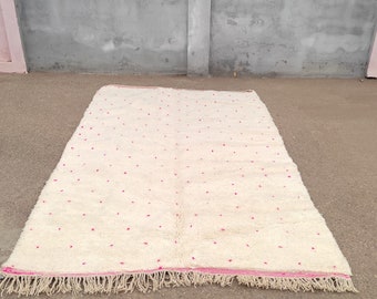 May include: Cream-colored rectangular rug with a pattern of small, evenly spaced red dots. The rug has a fringe along the bottom edge and a pink border. The rug is made of a soft, textured material.