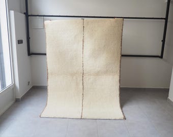 Puede incluir: Una alfombra grande, rectangular, de color crema con una textura shaggy. La alfombra tiene una costura vertical en el centro y un borde marrón. La alfombra está colgada contra una pared blanca, mostrando su tamaño y textura. Es un artículo de decoración del hogar.