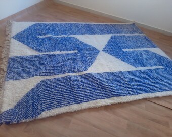 Puede incluir: Una alfombra blanca y azul con un patrón geométrico. La alfombra tiene un borde de flecos y está hecha de un material suave y esponjoso.