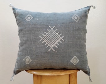 May include: A gray throw pillow with white embroidered diamond patterns. The pillow has tassels on the corners and is sitting on a wooden stool.