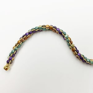 Può includere: Un braccialetto multi-colore con perline dorate, viola e verdi. Il braccialetto è realizzato con piccole perline quadrate.