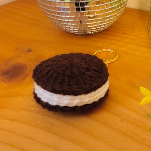 May include: A round, crocheted cookie keychain featuring two dark brown textured layers with a thick white center layer, attached to a gold-toned metal split ring. The item is crafted from yarn and mimics the appearance of a sandwich cookie.