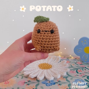 May include: A small, brown, crocheted potato plushie with a single green leaf on top and two black bead eyes. The word POTATO is printed in white letters at the top, flanked by two small white flower icons. The item is held against a floral background.