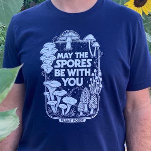 May include: Navy blue t-shirt with a white graphic of various mushrooms and the text "MAY THE SPORES BE WITH YOU" and "PLANT POSSE". The person is wearing a blue cap and standing in front of sunflowers.