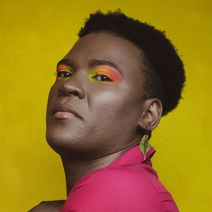 Puede incluir: Una persona con el pelo corto y oscuro que lleva una camisa rosa brillante y maquillaje de ojos colorido. Lleva un pendiente en forma de hoja verde.