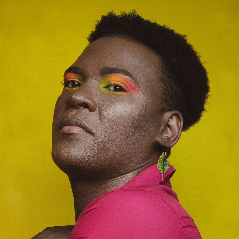 Puede incluir: Una persona con el pelo corto y oscuro que lleva una camisa rosa brillante y maquillaje de ojos colorido. Lleva un pendiente en forma de hoja verde.