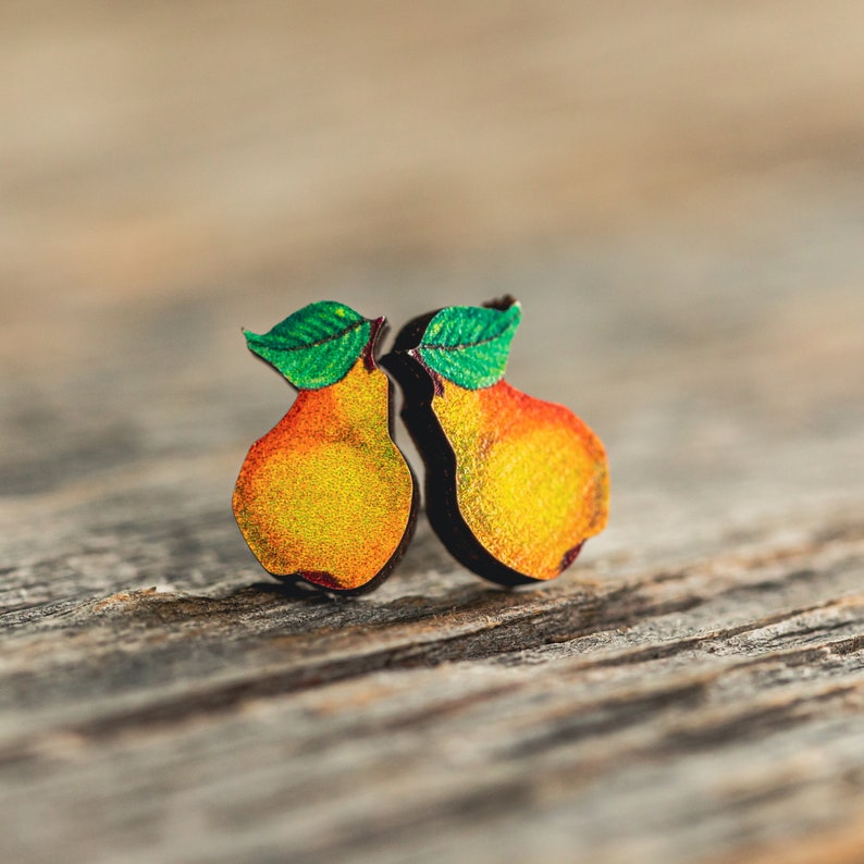 Small stud earrings with an original painting of a yellow and orange colored pear printed on die-cut wood. The two earrings are symmetrical to one another.