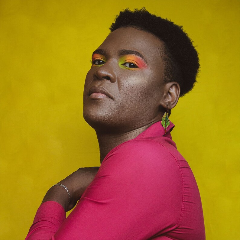 Puede incluir: Una persona con el pelo corto y oscuro que lleva una camisa rosa brillante y un pendiente de hoja verde. La persona tiene un maquillaje de ojos colorido y mira directamente a la c&aacute;mara.