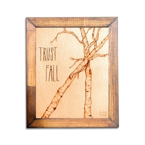 May include: A framed print with a watercolor illustration of two birch trees leaning against each other. The trees are brown and the background is a light orange. The text "TRUST FALL" is written in brown lettering on the print.