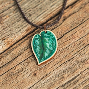 May include: A green leaf pendant necklace with a brown cord. The leaf is painted with a green and white pattern and is shaped like a heart.