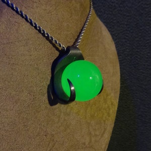Puede incluir: Un collar de cadena de plata con un colgante que presenta un orbe verde brillante encerrado en un diseño de garra de plata.
