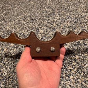 May include: A wooden cue rack with six slots for holding pool cues. The rack is brown and has two silver screws on the back.