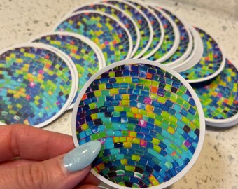 Op de afbeelding: Een stapel ronde stickers met een kleurrijk mozaïekontwerp. De stickers hebben een blauw, groen, geel en oranje mozaïekpatroon.