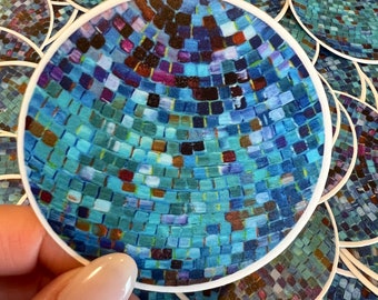 Op de afbeelding: Een close-up van een blauwe en groene discobal sticker. De sticker is rond en heeft een witte rand. De discobal is gemaakt van veel kleine vierkanten.