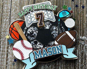 May include: A wooden cake topper with a black and white soccer ball, a baseball, a basketball, a football, a golf club and ball, and a green and white net. The topper also has a blue banner with the text "MASON" and a green banner with the text "HAPPY BIRTHDAY 7".