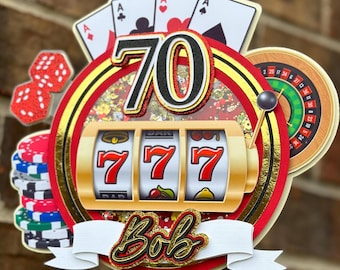 May include: A casino-themed birthday cake topper with a gold and red color scheme. The design includes a slot machine with triple sevens, playing cards, dice, and a roulette wheel. The number 70 is prominently displayed, along with the name "Bob".