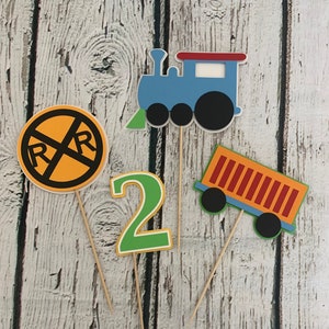 May include: Four cake toppers featuring a blue train, a yellow railroad crossing sign, a green number two, and an orange boxcar. The toppers are on wooden sticks.