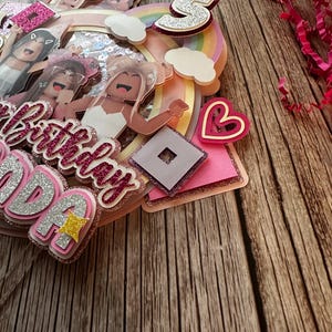 May include: A pink and white cake topper with a rainbow, clouds, a heart, a Roblox logo, and the words "Happy Birthday" and "Ava". The topper features several cartoon girls with pink and white clothing.