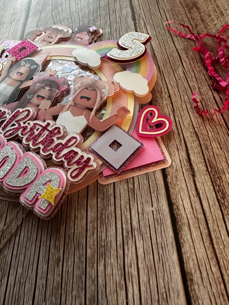 May include: A pink and white cake topper with a rainbow, clouds, a heart, a Roblox logo, and the words "Happy Birthday" and "Ava". The topper features several cartoon girls with pink and white clothing.