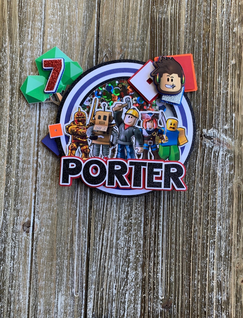 Pu&ograve; includere: Un topper per torta a tema Roblox con un cerchio blu, glitter e vari personaggi di Roblox. Il nome "PORTER" &egrave; scritto in lettere nere con un contorno rosso. Il numero "7" &egrave; su un blocco verde e rosso.
