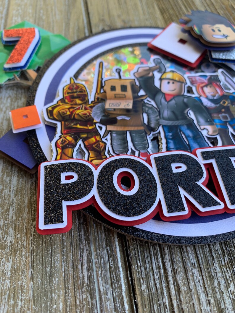 Pu&ograve; includere: Un topper per torta bianco e nero con la parola "PORTAL" in grandi lettere scintillanti. Il topper presenta vari personaggi di Roblox, tra cui un cavaliere, un robot, un operaio edile e un pirata.