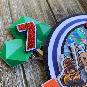 Pu&ograve; includere: Un numero sette rosso con glitter, una forma geometrica verde e un cerchio blu e argento con glitter con un cavaliere e una spada. Il cerchio ha un bordo nero e uno sfondo bianco. C'&egrave; anche un piccolo quadrato arancione con un quadrato nero al centro.