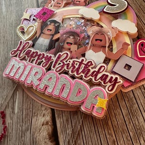 May include: A pink and silver cake topper with the words "Happy Birthday Miranda" and a rainbow with clouds. There are also several cartoon figures in the design.