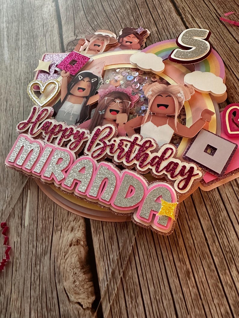 May include: A pink and silver cake topper with the words "Happy Birthday Miranda" and a rainbow with clouds. There are also several cartoon figures in the design.