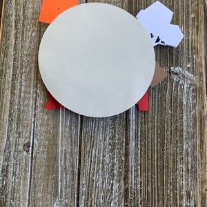 Pu&ograve; includere: Un cerchio di carta bianco con forme di carta rossa, arancione e bianca attaccate ad esso. Il cerchio &egrave; attaccato a due bastoncini di plastica trasparente.