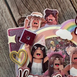 May include: A pink, purple and yellow cake topper with a rainbow, a cloud and four cartoon figures. The figures are holding a red and pink block with a star on top. The cake topper says "Happy Birthday".