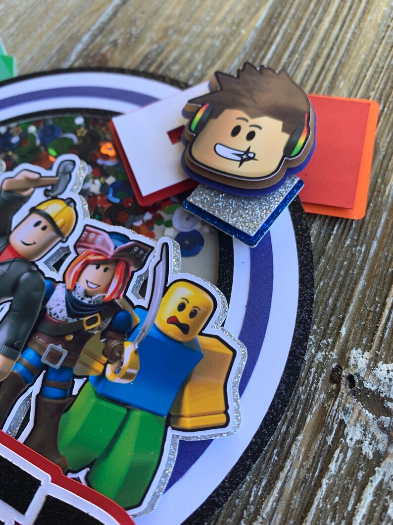 Pu&ograve; includere: Un topper per torta colorato con diversi personaggi di Roblox, tra cui un personaggio con una camicia blu e una testa gialla, un personaggio con un cappello da pirata rosso e una camicia blu e un personaggio con una camicia blu e una testa gialla. I personaggi sono in piedi su un cerchio bianco con un bordo di glitter argentato. Il topper per torta include anche un quadrato rosso, bianco e blu, un quadrato di glitter argentato e un personaggio di Roblox con capelli castani e cuffie.