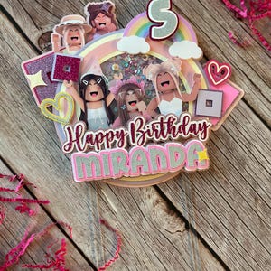 May include: A pink and white cake topper with a rainbow, clouds, and four cartoon figures. The topper says "Happy Birthday Miranda" and has a star and a heart.
