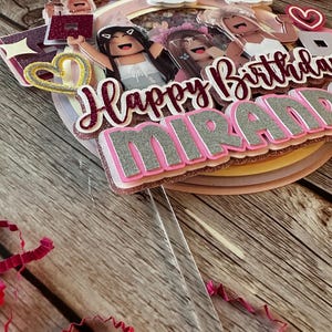 May include: A pink, white and gold cake topper with the words "Happy Birthday Miranda" and a rainbow. The topper features several cartoon figures with pink and white clothing.