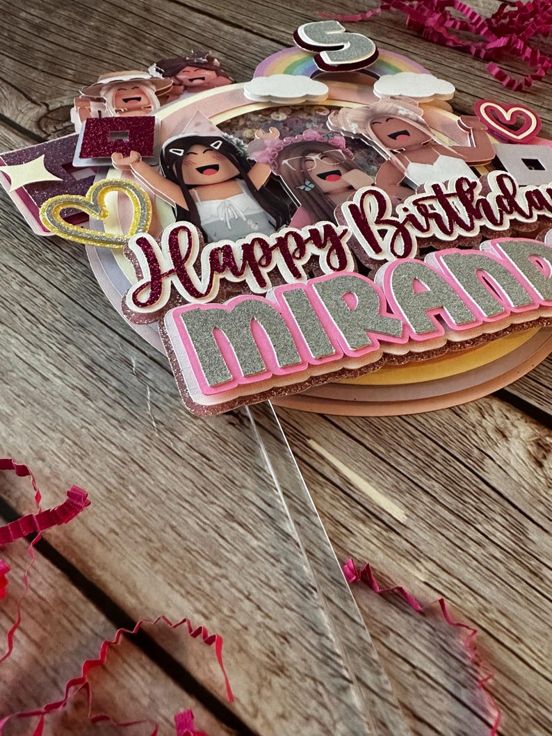 May include: A pink, white and gold cake topper with the words "Happy Birthday Miranda" and a rainbow. The topper features several cartoon figures with pink and white clothing.