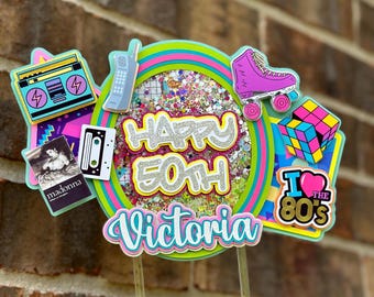 May include: A cake topper with a 1980s theme. It features a colorful circle with the text "Happy 50th Victoria" and various 80s icons like a boombox, cassette tape, roller skate, and a Rubik's Cube. The text "I love the 80s" is also included.