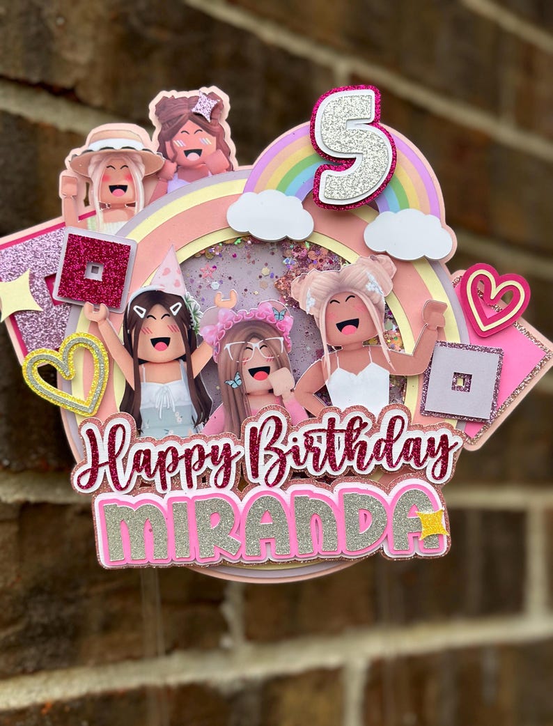 May include: A pink and white cake topper featuring a rainbow, clouds, and four cartoon girls. The topper reads "Happy Birthday Miranda" and has a pink number 5 on top.