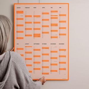 Könnte beinhalten: Ein Wandkalender mit hellpfirsichfarbenem Hintergrund und einem leuchtend orangefarbenen Rand. Der Kalender zeigt zwölf Monate von Januar bis Dezember in schwarzer Schrift, wobei bestimmte Daten durch orangefarbene Balken markiert sind.