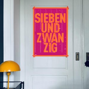 Könnte beinhalten: Ein rechteckiges Wandplakat mit einem leuchtend magentafarbenen Hintergrund und kräftiger orangefarbener Typografie, die SIEBEN UND ZWANZIG liest. Das Design enthält vertikale Kalenderspalten mit kleinem Text und das Jahr 2027.