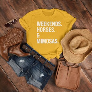 May include: A yellow t-shirt with the text "Weekends. Horses. & Mimosas." on it. The shirt is laid out on a wooden surface with a pair of brown cowboy boots, a brown leather purse, a straw hat, and denim shorts.