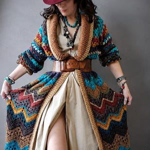 Könnte beinhalten: Ein bordeauxroter Filzhut mit schwarzem Band, eine lange Häkelstrickjacke mit mehrfarbigem Zickzackmuster in Türkis, Braun, Rot und Gelb, ein beiges Hemdkleid, ein breiter brauner Ledergürtel, mehrlagige Perlenketten und graue Socken.