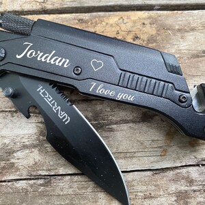 May include: Black multi-tool knife with a partially open blade. The knife has the name "Jordan", a heart symbol, and the phrase "I love you" engraved on it. The blade has the word "WARTECH" printed on it.