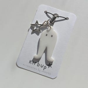 May include: A white acrylic charm shaped like a pair of legs with two small dots, attached to a black cord loop with a silver-toned star charm. The charm is mounted on a white display card printed with the text kirbup accessories by unkirbb.