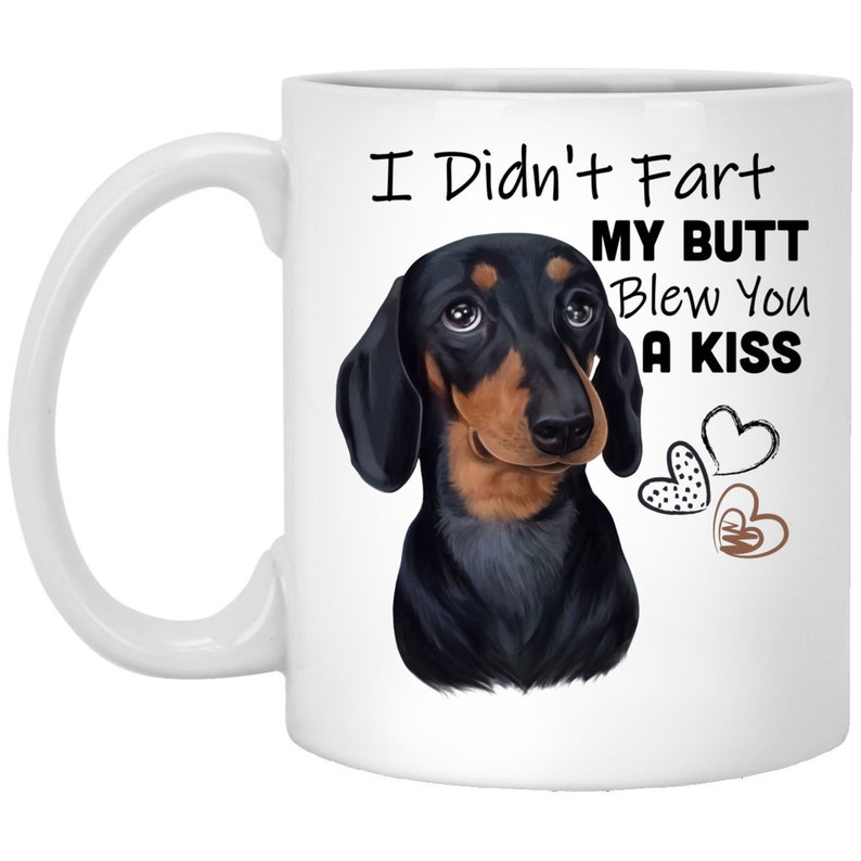 May include: White ceramic coffee mug featuring a black and tan dachshund illustration. The text on the mug reads "I Didn't Fart My Butt Blew You A Kiss" with three hearts.
