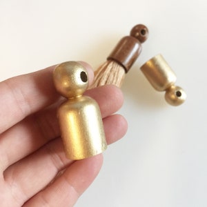 Puede incluir: Primer plano de componentes de borlas de madera pintadas de dorado. Una pieza se sostiene en una mano, mientras que otras est&aacute;n dispersas cerca. Las borlas son una mezcla de dorado y marr&oacute;n, con un cepillo de fibra natural.