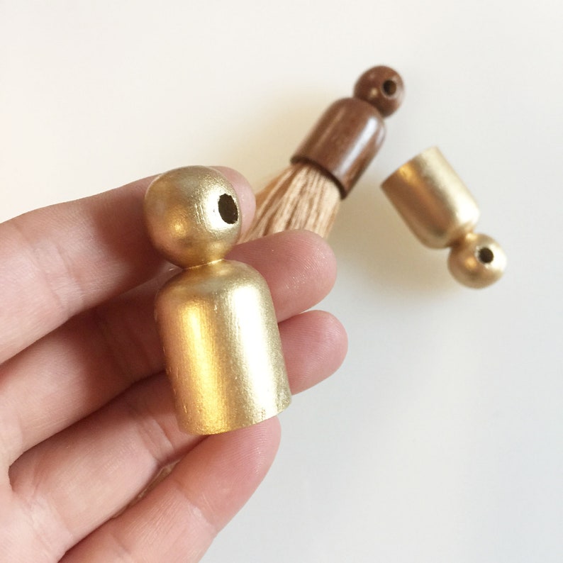 Puede incluir: Primer plano de componentes de borlas de madera pintadas de dorado. Una pieza se sostiene en una mano, mientras que otras est&aacute;n dispersas cerca. Las borlas son una mezcla de dorado y marr&oacute;n, con un cepillo de fibra natural.
